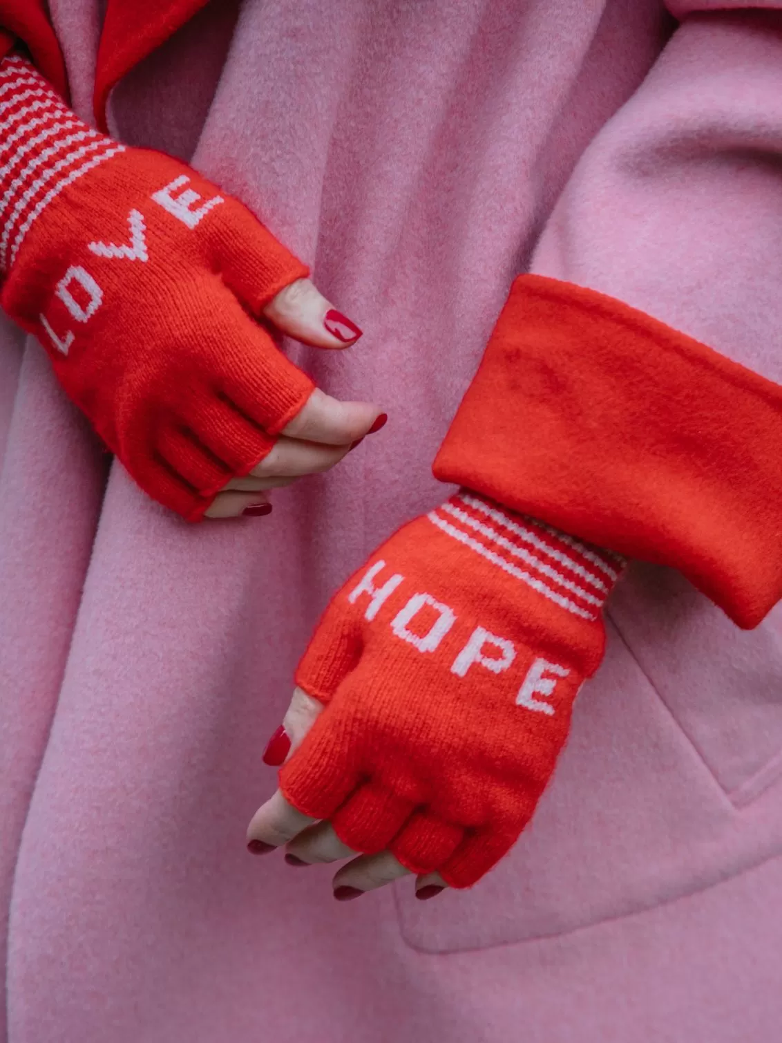 A classic red fingerless knitted glove with the words LOVE and HOPE in dusky pink and stripe cuff with a bubblegum pink edge detail. Made from 100% Lambswool with Wool spun in Yorkshire. 