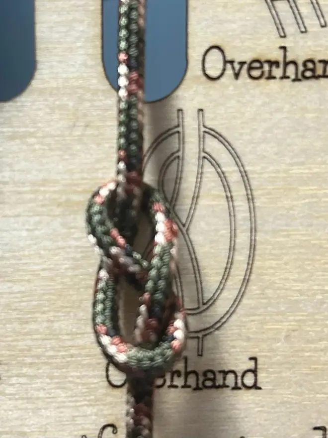 Wooden knot training board with engraved knot diagrams and red and black ropes for practice, displayed outdoors among pinecones and leaves.