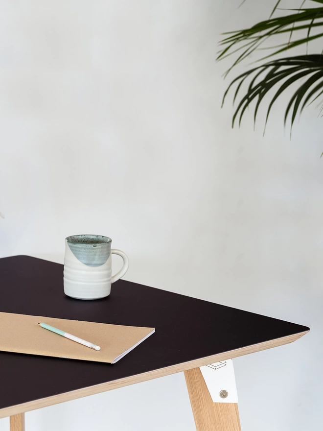 corner of a stylish and minimalist desk with black Fenix top, white coloured steel brackets and solid oak legs, with a mug and notebook on it.