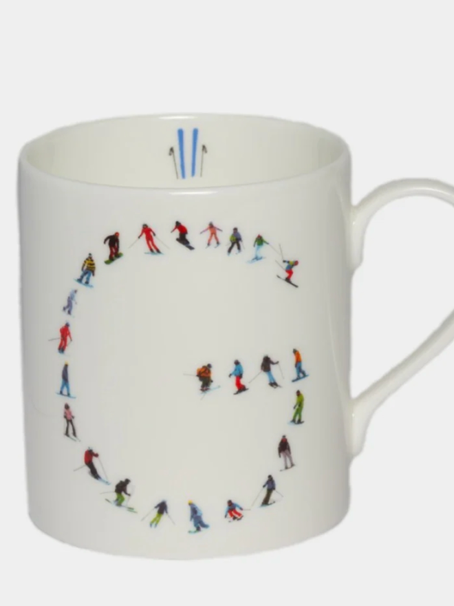 A simple white mug illustrated with a ‘G’ made of small, multicoloured skiers. The mug is set against a plain, white background.