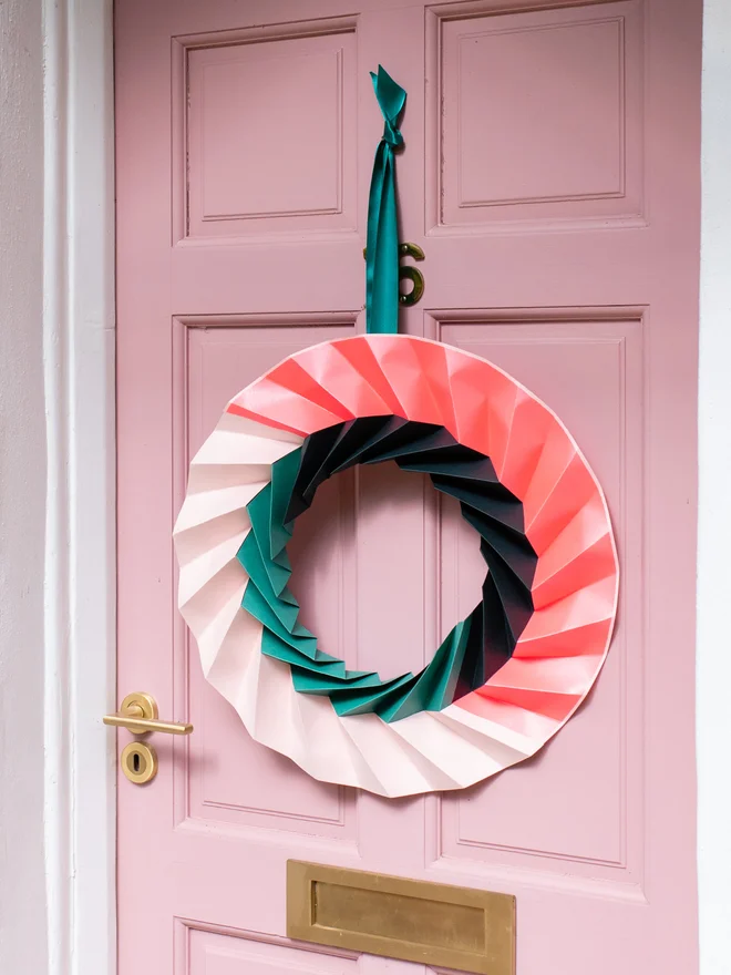 Modern origami wreath hanging on a pink front door. The wreath is neon pink, pale pink, and green in colour.