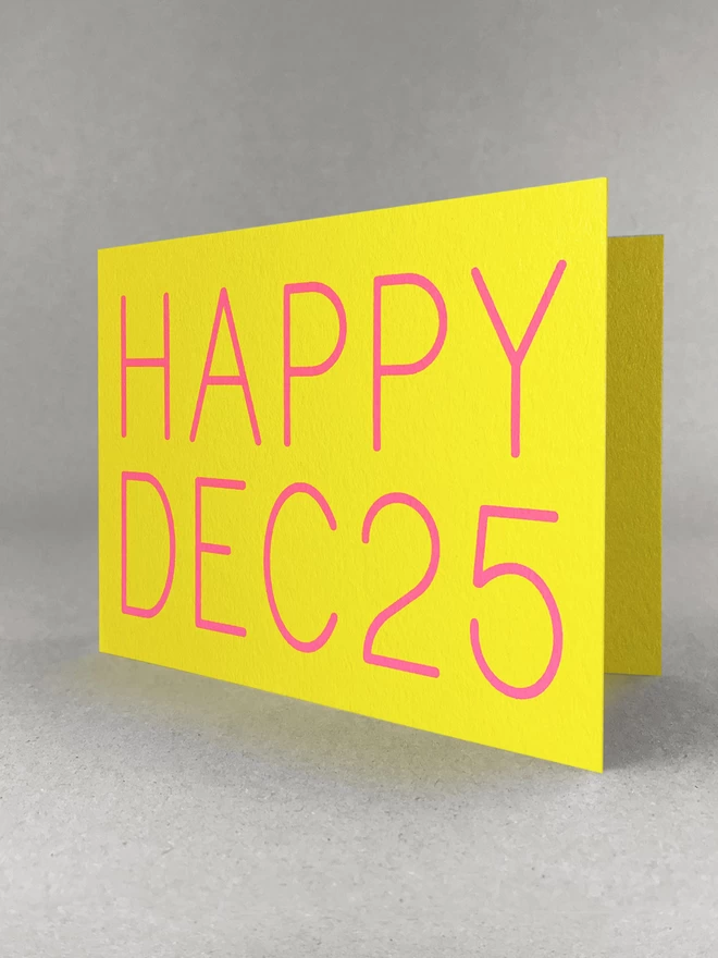 Industrial yellow card with neon pink HAPPY DEC 25 screenprinted upon. Stood on a pale grey backdrop, slightly open.