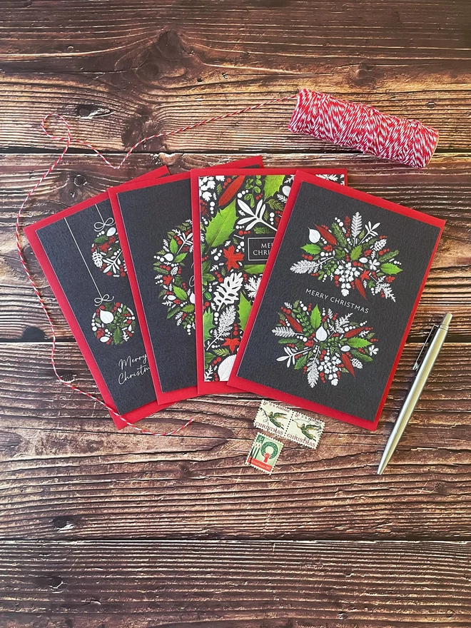 Pack of 4 Christmas Cards with Red, White, and Green Pressed Winter Leaf Designs - Arranged on Wooden Tabletop - Twine Spool, Christmas Postage Stamps, Silver Pen