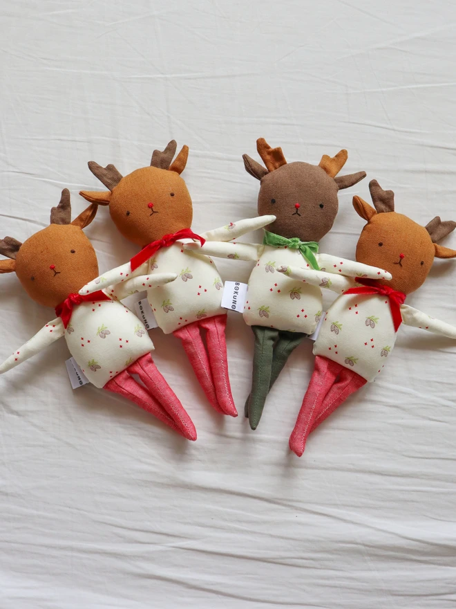 Four reindeer dolls arranged on white sheets. Each doll has soft antlers, embroidered facial features, and a festive pinecone print outfit with either a red or green velvet bow. Two have green legs, and two have sparkly red legs. The dolls have "BOKUNO" fabric tags.