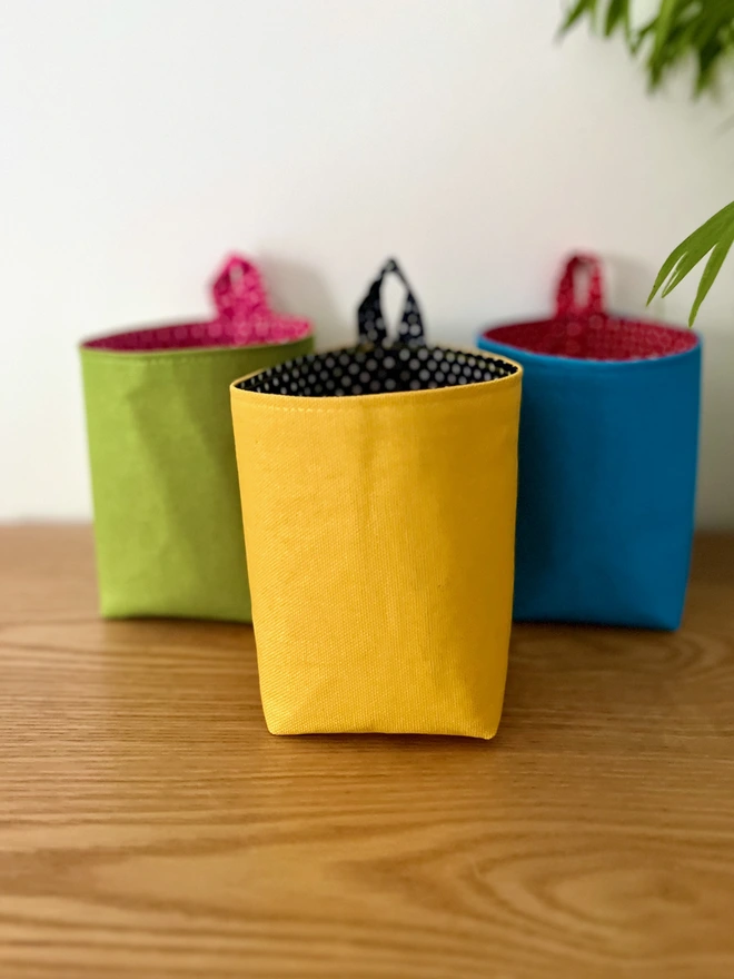 3 small hanging fabric bags in blue, green and yellow colours on a wooden surface.