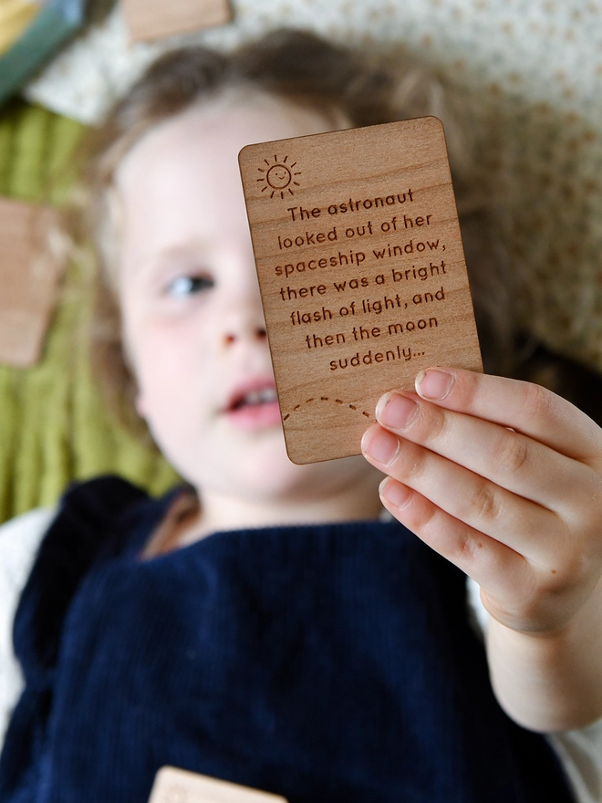 A young girl wearing blue dungarees lays down and is reading a wooden story card that she holds above her head.