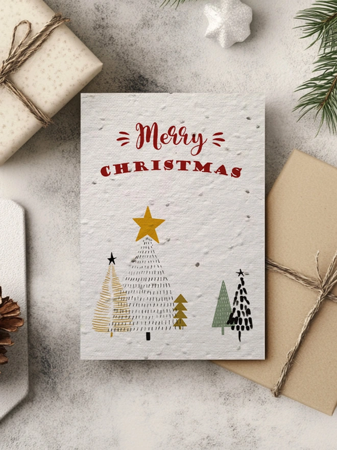Christmas Card featuring Christmas trees and 'Merry Christmas' across the top. In the background is presents, pine cones and greenery.