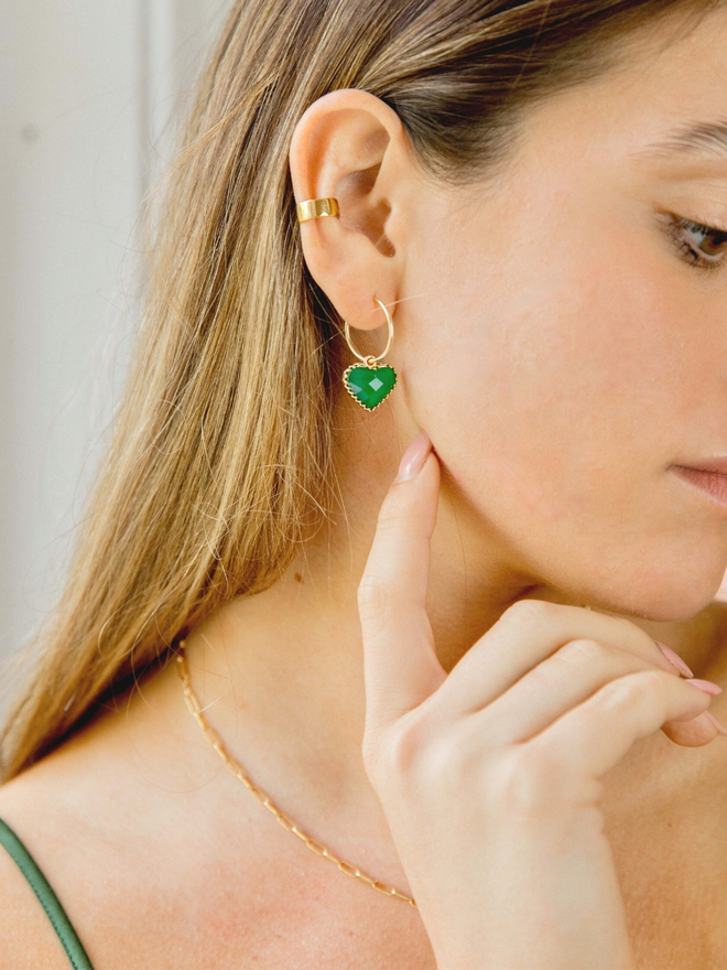 Woman wearing a small gold hoop earring with a green faceted heart charm hanging from it