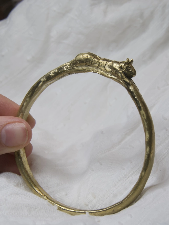 Image of a gold toned bangle being held on a white cloth, with a hand carved tiger stretched across the top of the bangle 