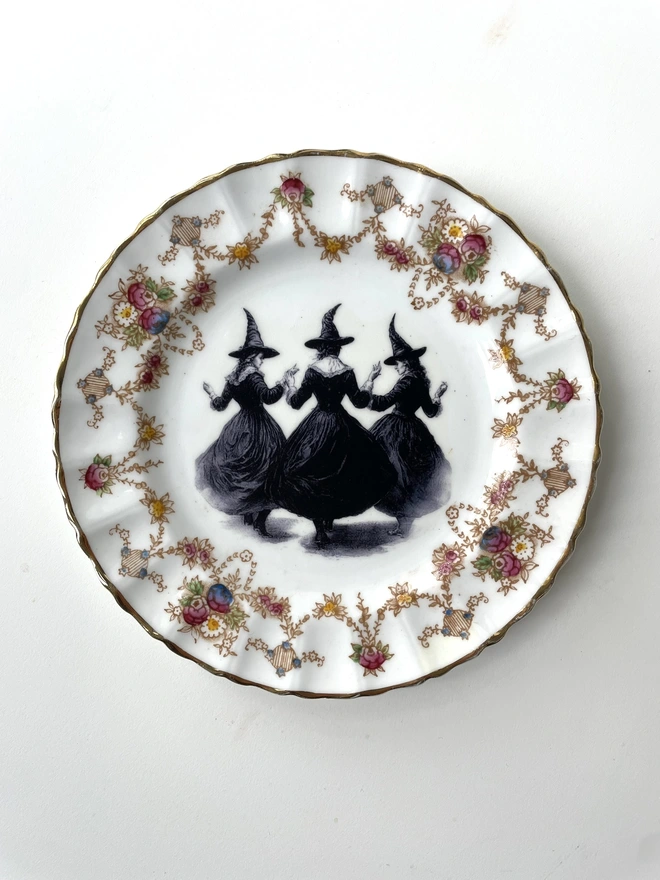 a coven of 3 witches dance hand in hand, the image is victorian in black and white and is printed directly onto an ornate vintage plate