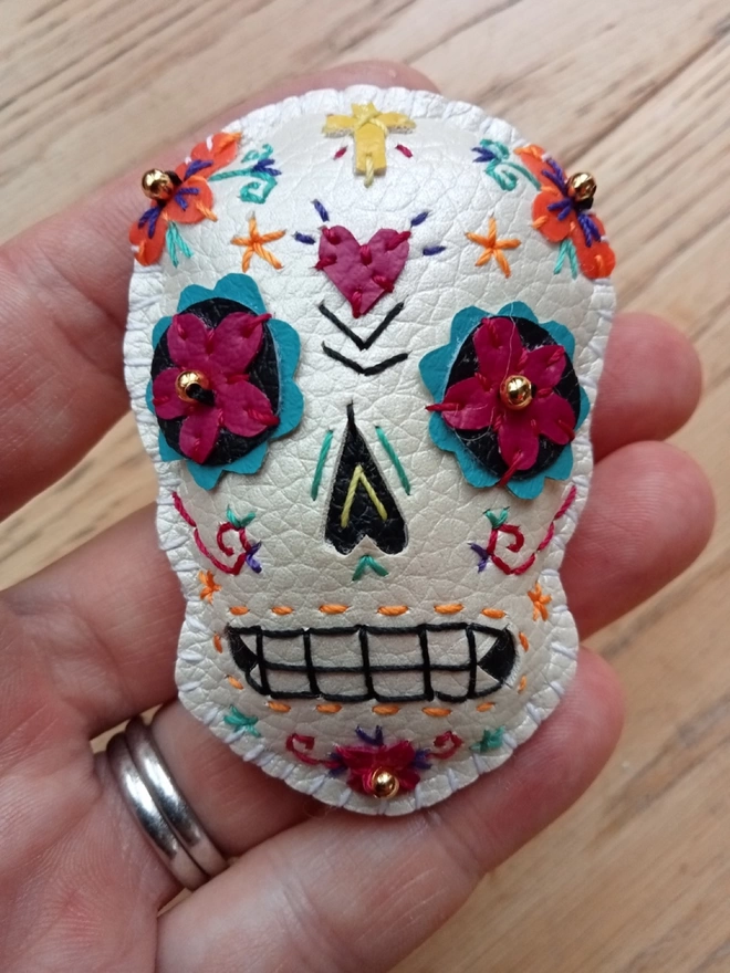 A hand stitched faux leather white Mexican Day of the Dead inspired sugar skull brooch with  colourful stitching and gold bead details, held by a hand, on a wooden background 