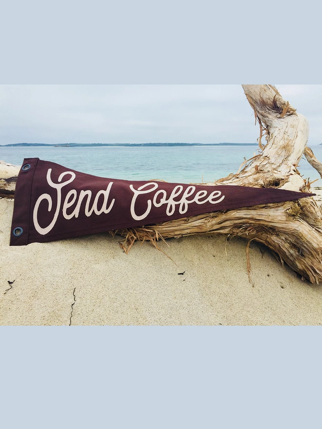 A photo of an espresso brown pennant flag with the words Send Coffe in a latte coloured canvas. The pennant is resting on a piece of wood that has washed up on a sandy beach. 