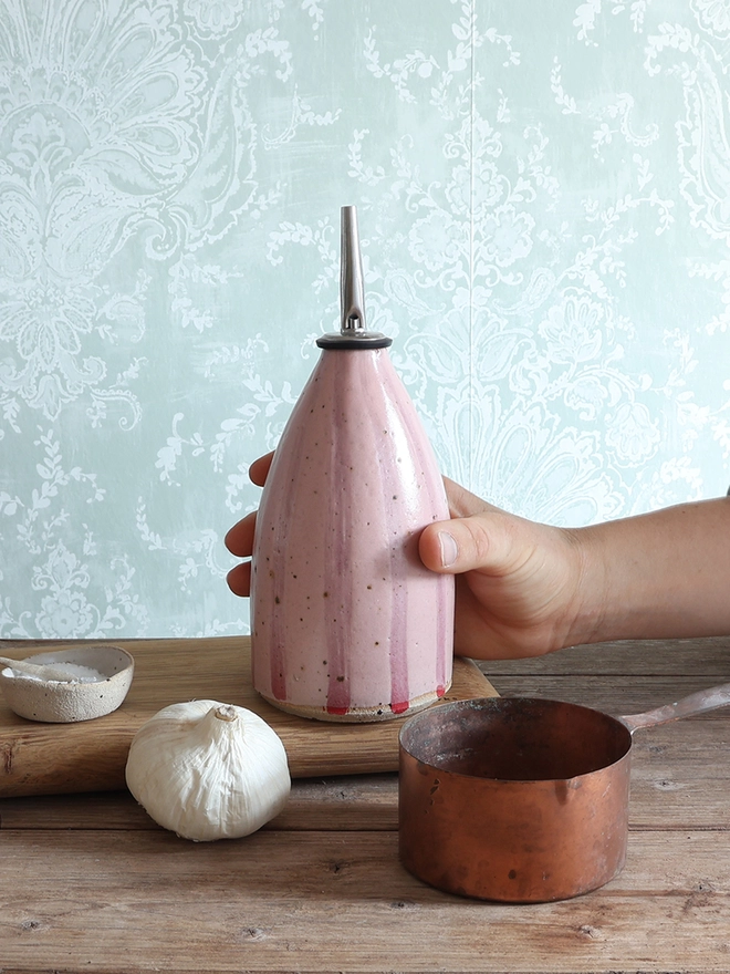 A female hand holds a Handmade Ceramic Stoneware Oil Pourer in Pink Stripe above a wooden surface. The oil pourer bottle is surrounded by bulb of garlic, a brass measuring cup, a wooden chopping board and small ceramic dishes containing salt and pepper. 