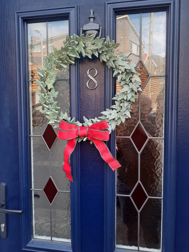 Handmade verdigris and copper ceramic ivy wreath hanging on a dark blue front door, finished with a festive red velvet bow.