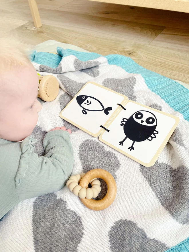 A baby lies on the floor having 'tummy time' on a knitted balnketr with a grey and white cloud design and mint green edging. The baby is staring at a black and white board book  and a wooden teething ring lies beside it.