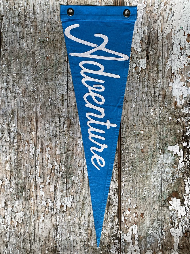 A nautical blue canvas pennant flag hanging vertically on a wooden wall with white canvas lettering saying Adventure.