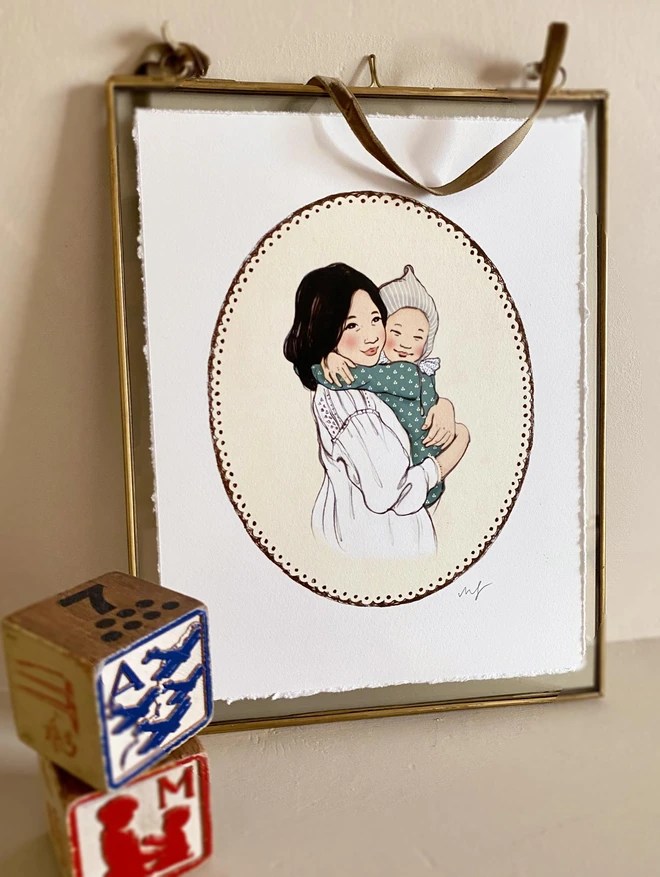 a phot of a mummy and me art print displayed in a glass frame on a mantle piece. it is an asian mother and baby cuddling hand drawn in a vintage story book style