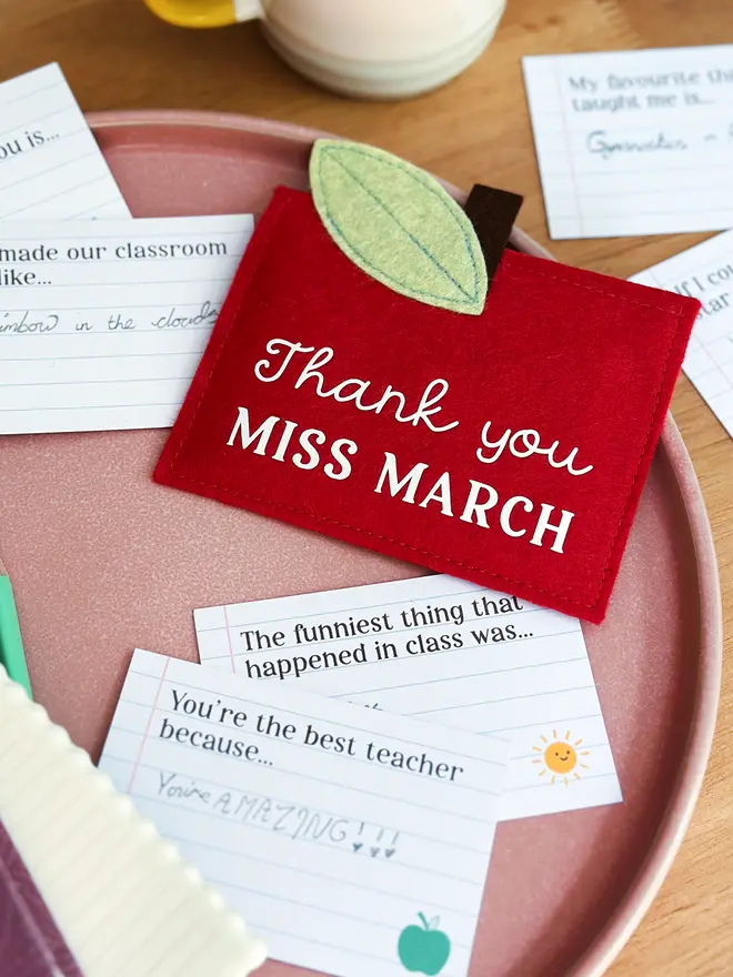 A handmade red felt apple-shaped envelope with "Thank you Miss March" written on the front. The heirloom apple envelope surrounded by completed prompt cards with handwritten messages from a child, displayed on a pink plate and wooden desk. It's a thoughtful personalised gift for teachers from children.
