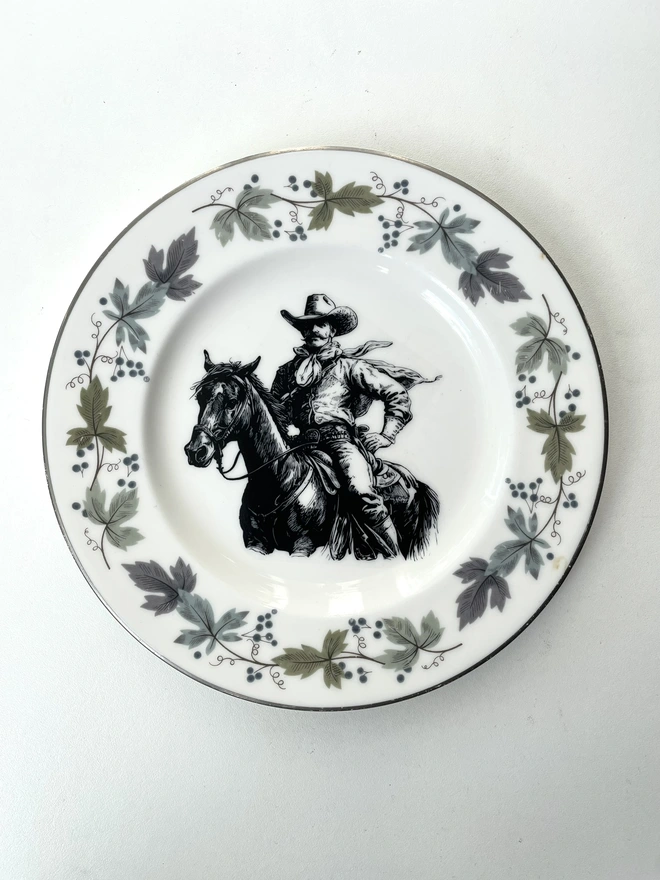 vintage plate hanging on a white wall, it has a decorative border, with a printed vintage illustration of a cowboy in the middle 