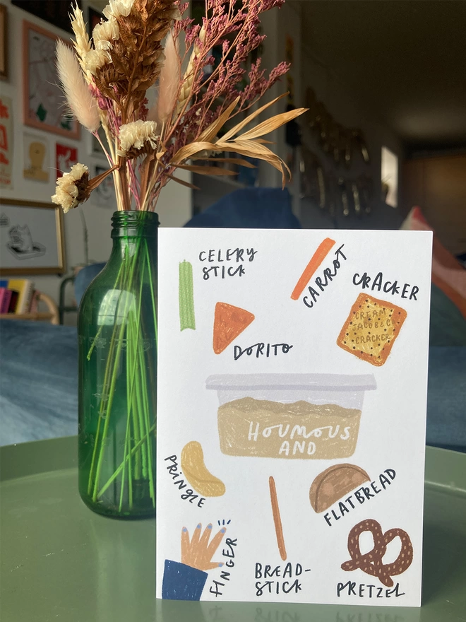 Greetings Card illustrated with houmous and snacks to dip. Placed on a table with flowers in the background.