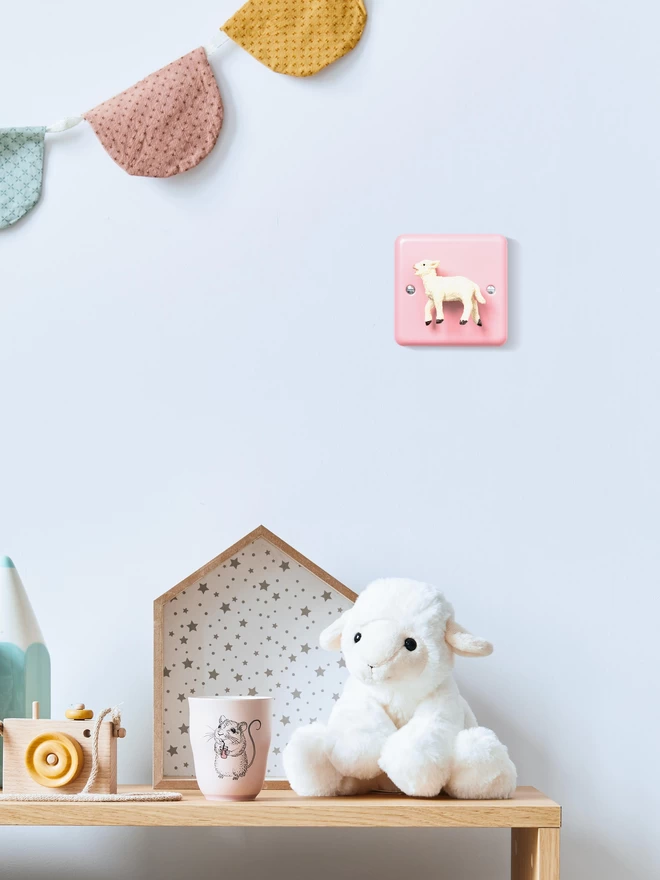 A pink dimmer light switch with a lamb as the rotary knob sits on a white wall. In the foreground there is a low wooden table with a soft lamb toy, a cream mug, a wooden toy camera and a house shaped storage box. There is pastel bunting on the wall. The brand of the light switch is Candy Queen Designs.