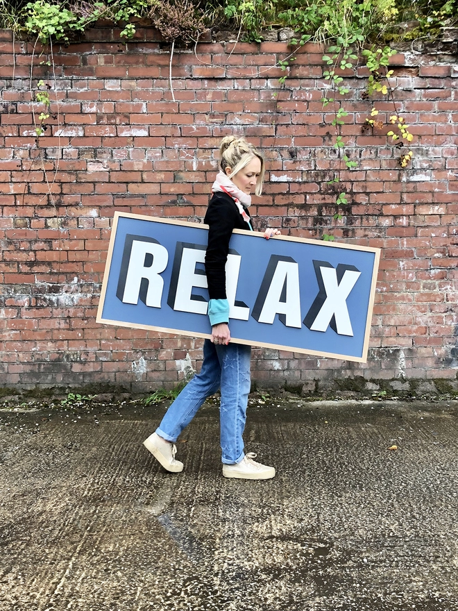 A person standing in front of a red brick wall holding a painted wooden sign which reads RELAX on a navy painted back board 
