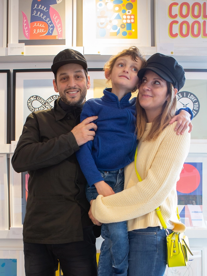 A Dad, young son and Mum stand in front of a wall of brightly coloured art prints. They are smiling.
