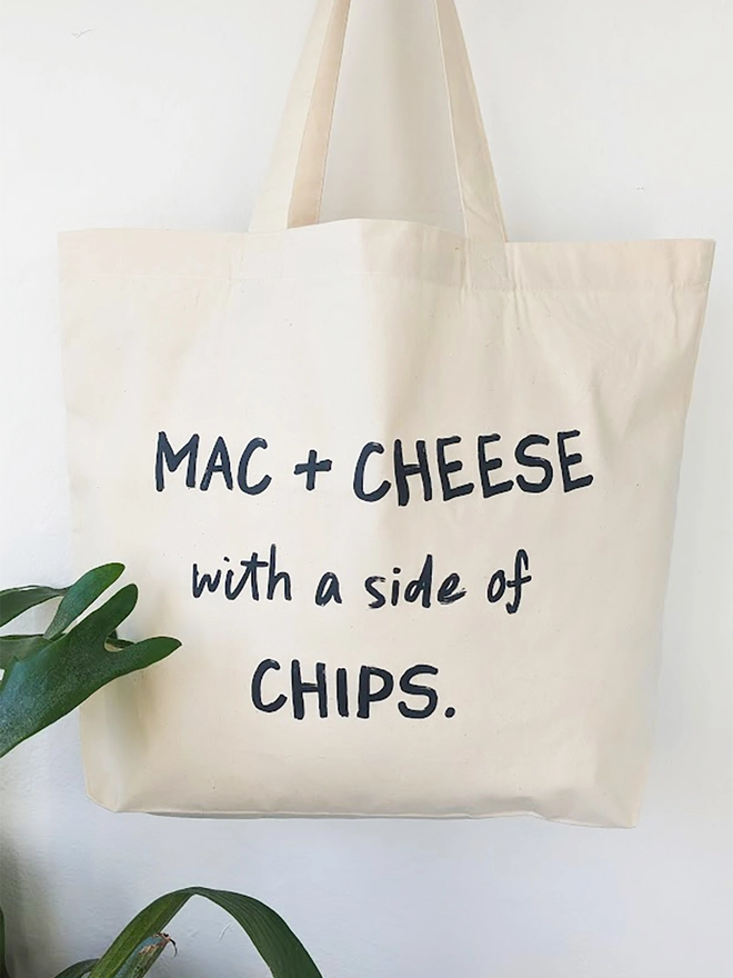 A natural coloured canvas tote with black slogan reading Mac and Cheese with a side of Chips