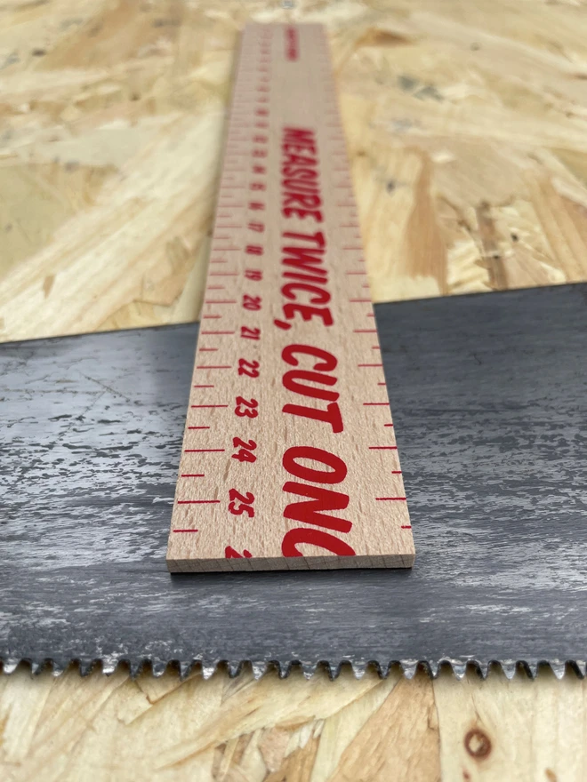 End view of a Measure Twice Cut Once typography on a wooden screenprinted ruler, showing the clean edge. Sat on a serrated saw and all on a chipboard background