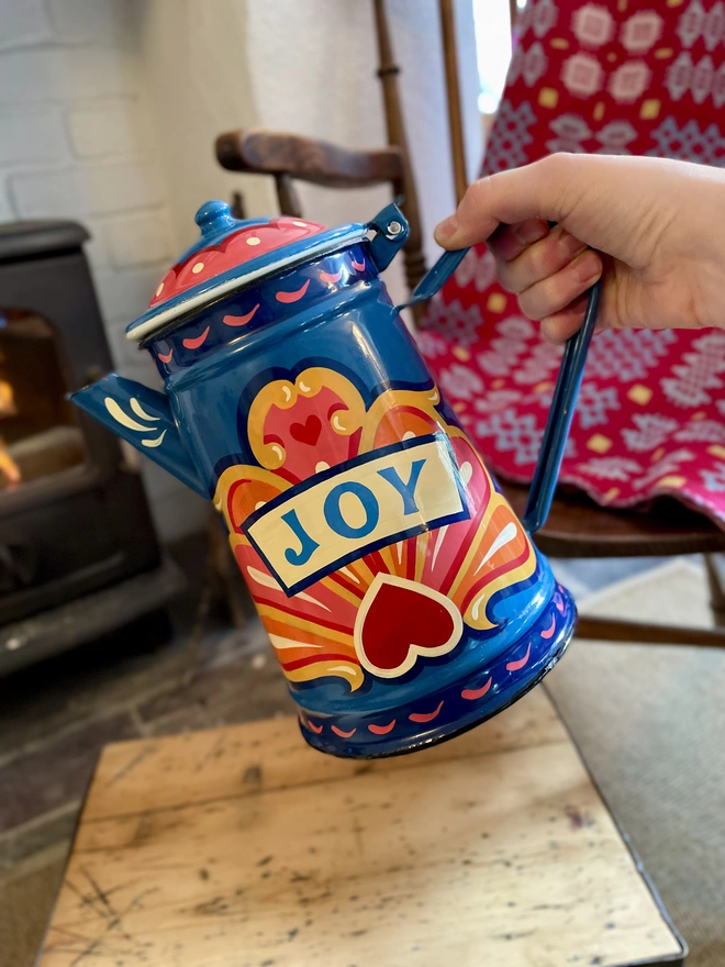 Blue vintage coffee pot, hand painted with a fairground inspired design in pink, orange and yellow. 'Joy' is painted in a banner through the centre of the design.