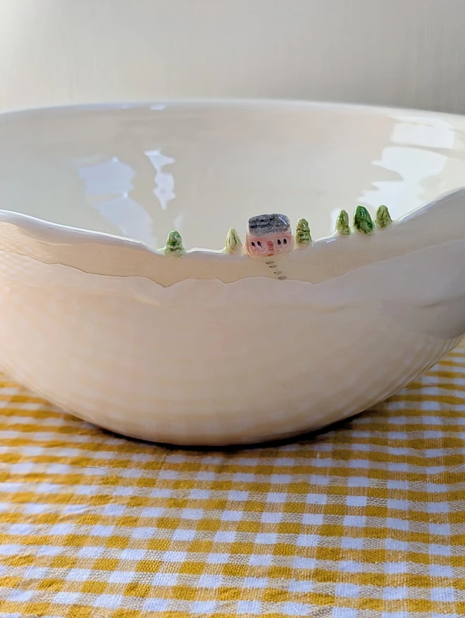 Handmade ceramic white bowl with a tiny pink house and tree details on the rim and white glaze on a yellow gingham table cloth