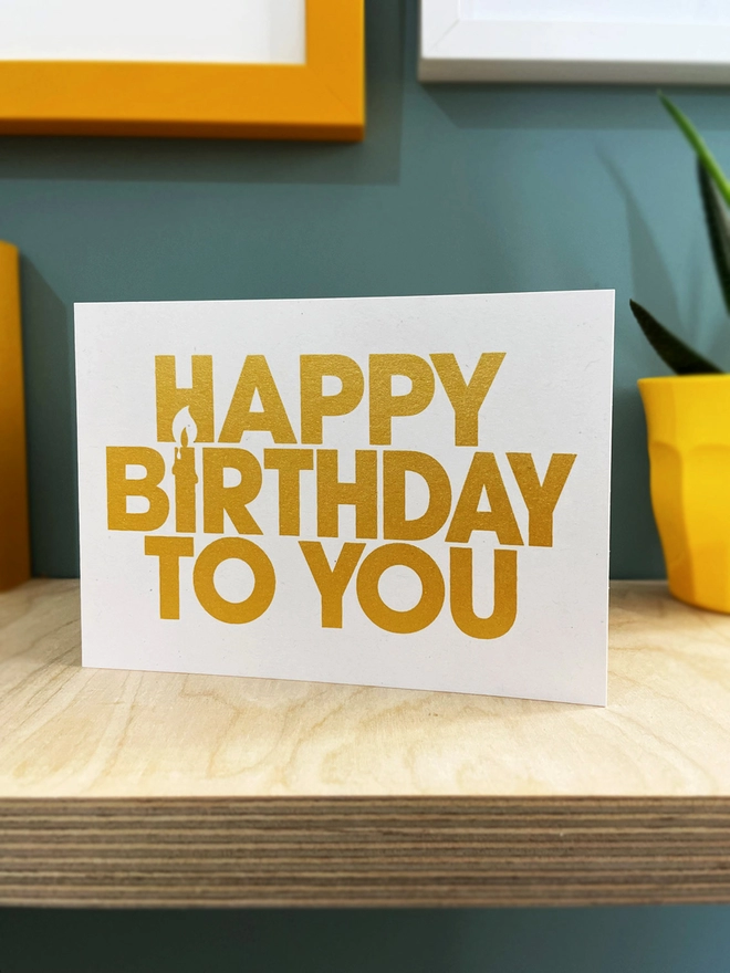 Happy Birthday screen printed in gold ink on a white landscape card, stood on a plywood shelf with hints of framed artwork around the edges , duck egg blue background.