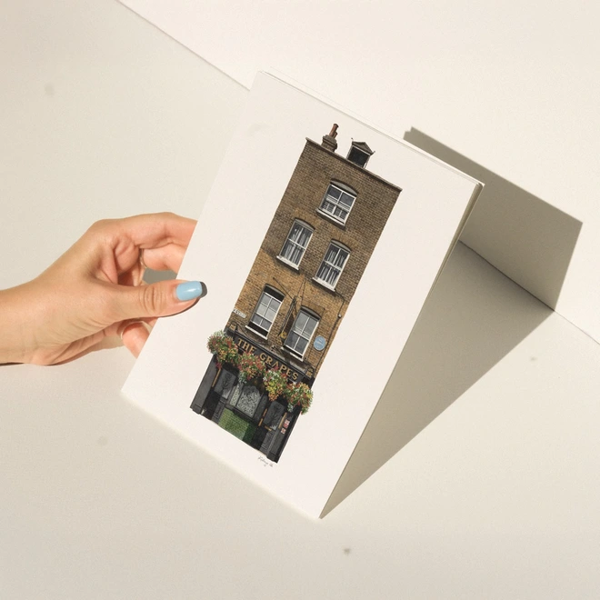 A hand with blue nail polish holding a print featuring an illustration of the tall, brick facade of The Grapes pub against a white background.