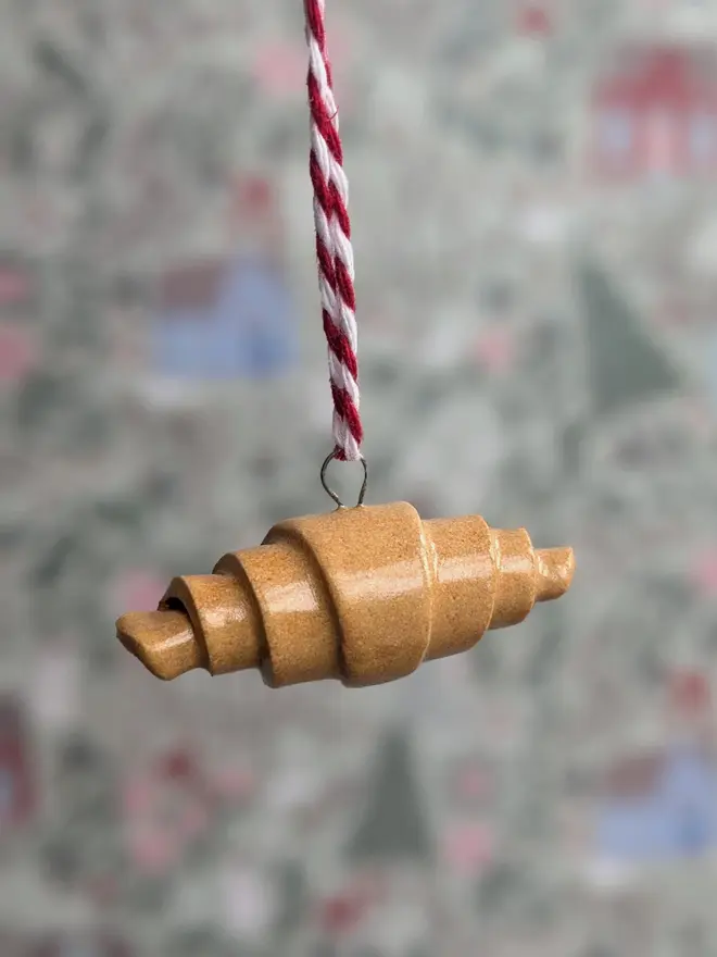 ceramic croissant hanging christmas decoration. a close up image of a small ceramic croissant hanging from red and white thread.