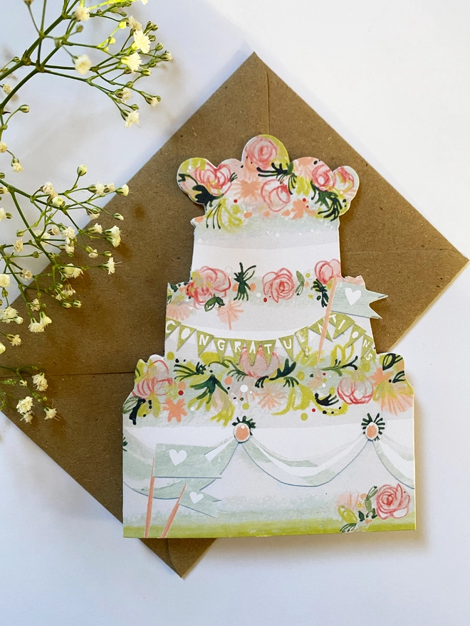 Esther kent illustrated greetings card in the shape of a tierd cake decorated with pink flowers and greenery.