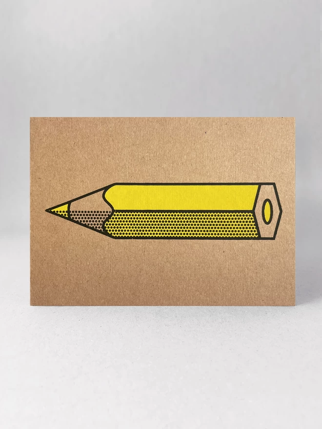 Kraft brown card, stood in a light grey studio background. The design is a yellow pencil, horizontal with a black outline and halftone detail. The card is flat to the camera.