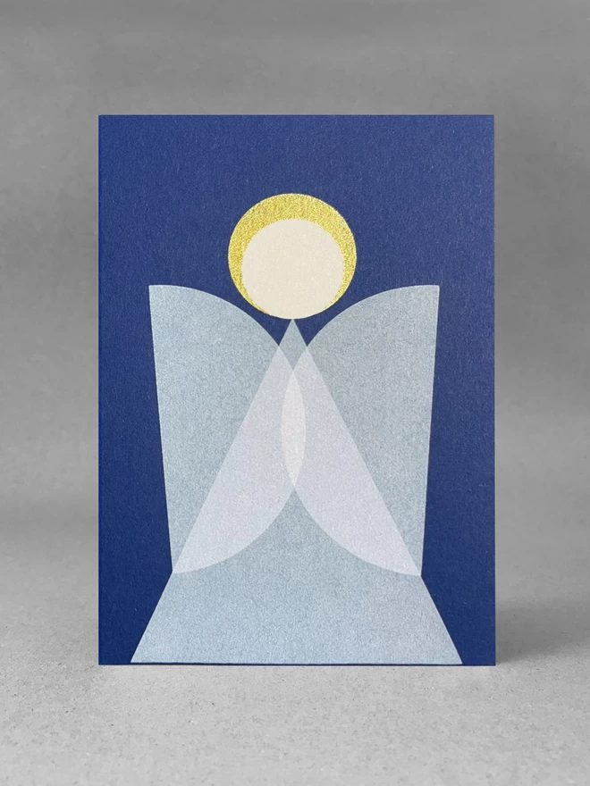 Geometric angel design, blue card, white overlapping wings and a gold halo. Sits in a light grey studio