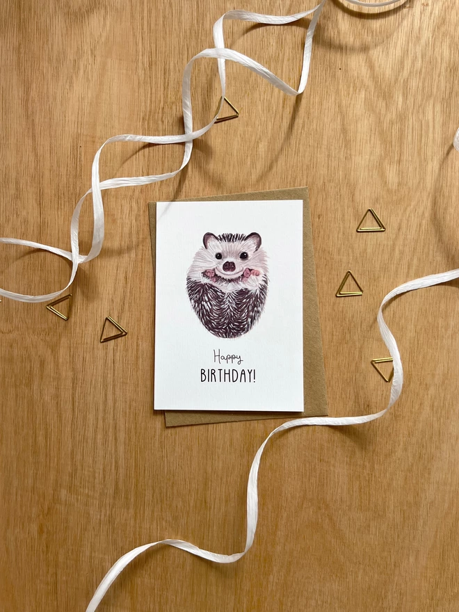 A greetings card with a white background featuring a baby hedgehog on their back with the phrase "happy birthday" underneath