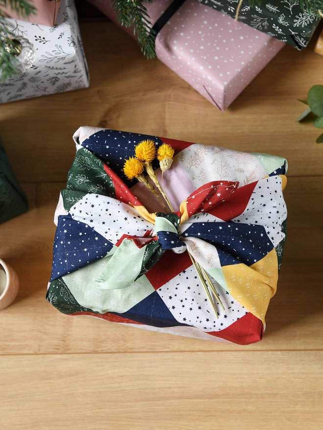 A gift wrapped in patchwork cotton fabric wrap, and a small posy of yellow flowers is being tucked into the knot.