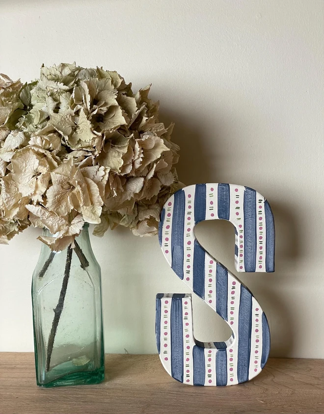 A hand-painted decorative initial letter  with stripes on a wooden table against a light wall. 