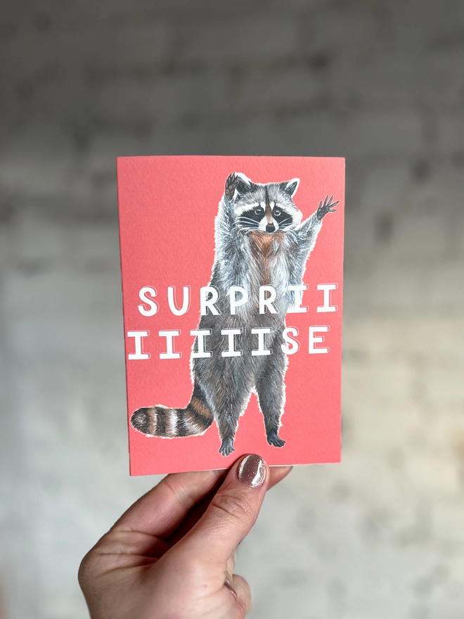 a bright coral pink greetings card featuring an illustration of a raccoon with its hands in the air with the text “surpriiiiise”