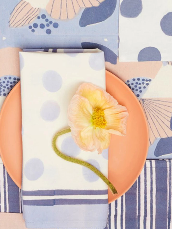 Close up of a block printed napkin in a pale blue spot design over a soft orange plate laid on a navy stripe and floral fabric 