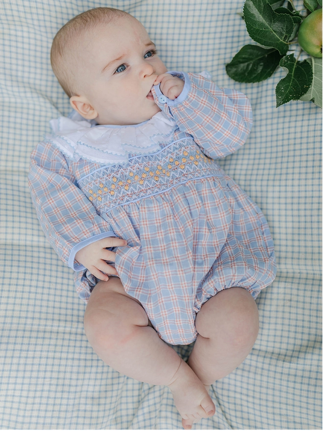 A baby wearing a check smocked romper with a white frill collar likes on a check sheet