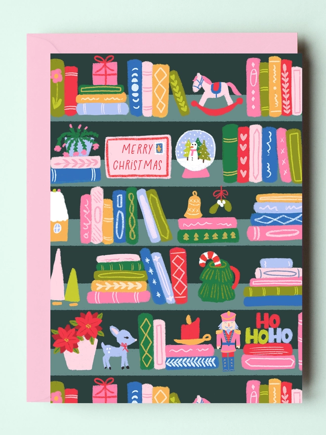 A Christmas bookshelf card laying flat with a pink envelope.