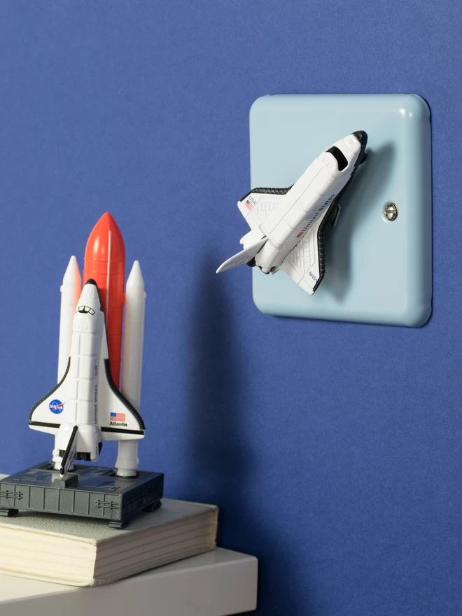 A pastel blue children's space themed dimmer light switch with a white space shuttle as the rotary knob to turn the lights on and off on a royal blue wall next to a white shelf with a toy space shuttle sitting on a book with a white cover. The light switch plate is pale blue and made of metal, epoxy coated steel by Varilight. The space rocket is made of metal. The children's light switch brand is Candy Queen Designs.