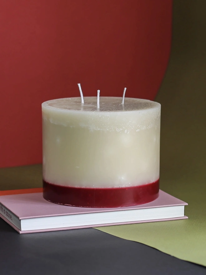 Oudh, Yuzu & Patchouli Candle (Set Of 1 Or 3 Wicks). the oudh, yuzu and patchouli candle sits on a pink book against a red and olive green background, showing its three wicks and cream wax with a deep red base. the two-tone colouring and textured, slightly uneven wax surface give the candle a rustic, hand-poured finish. the bold, contrasting background colours highlight the candle's warm tones.