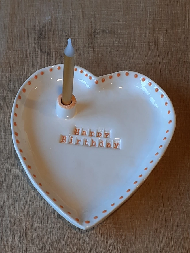 Handmade white heart-shaped ceramic Happy Birthday dish with orange lettering and dotted edging, shown with a birthday candle in the built-in holder.