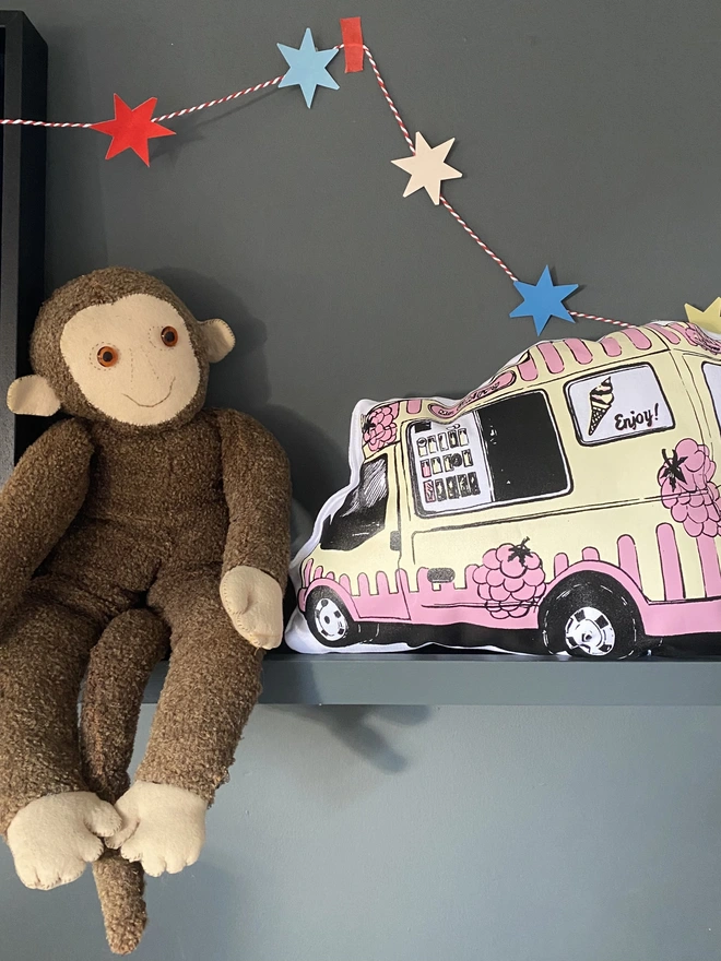 Ice cream van handmade cushion by the artist Flo Snook. A pink and cream cushion in the shape of an ice cream van on a shelf next to a monkey stuffed toy with a star garland hanging above