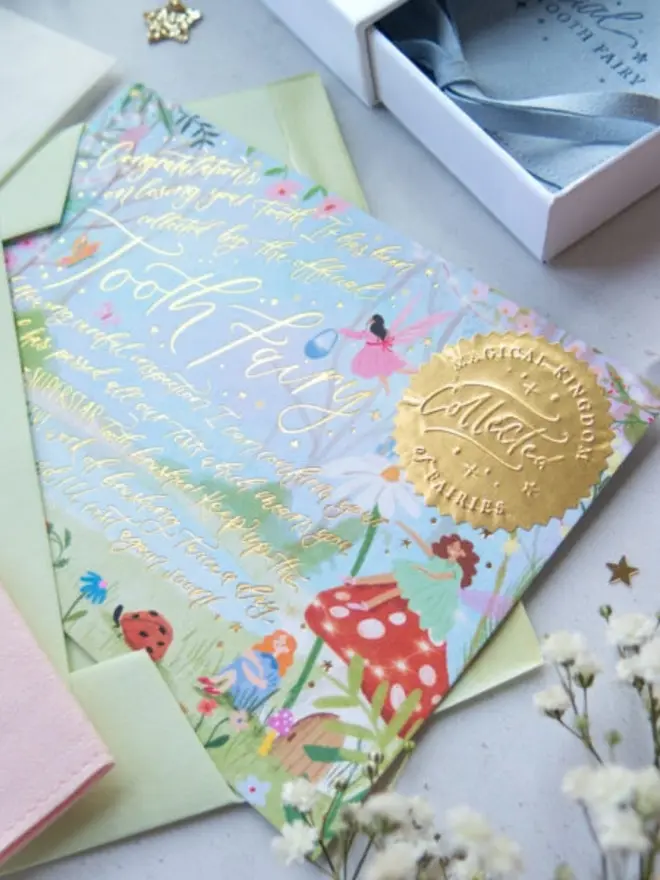 A close-up of the garden notecard's gold foil message congratulating a child on losing a tooth. An embossed gold "Magical Kingdom of Fairies Collected" seal sits beside a fairy on a red toadstool.