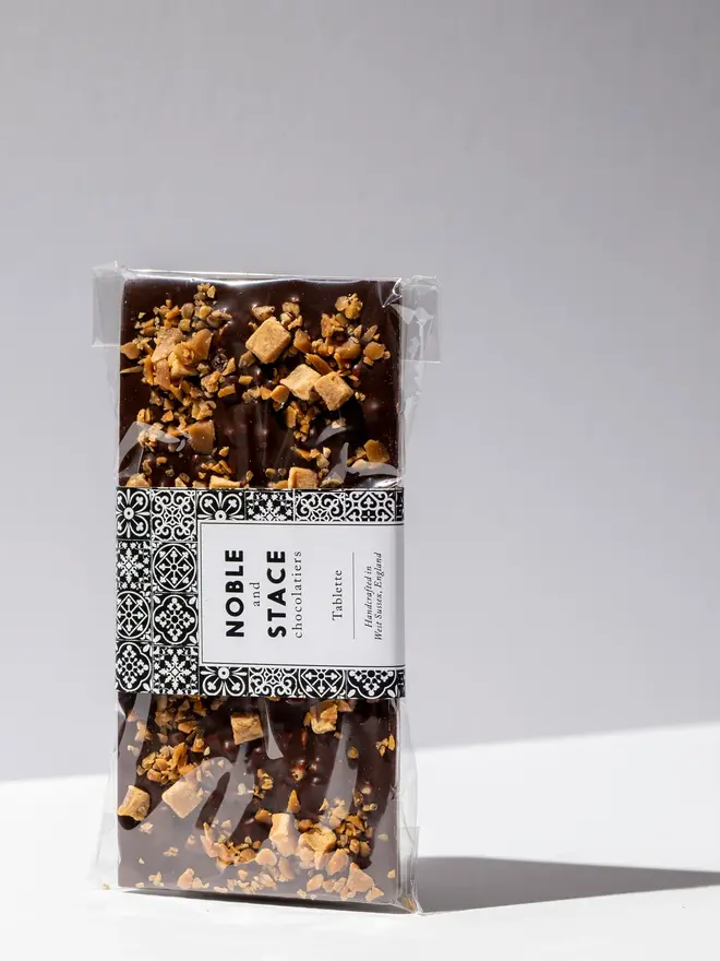 Honeycomb, toffee & fudge chocolate tablette wrapped in clear plastic and standing on a white table. 